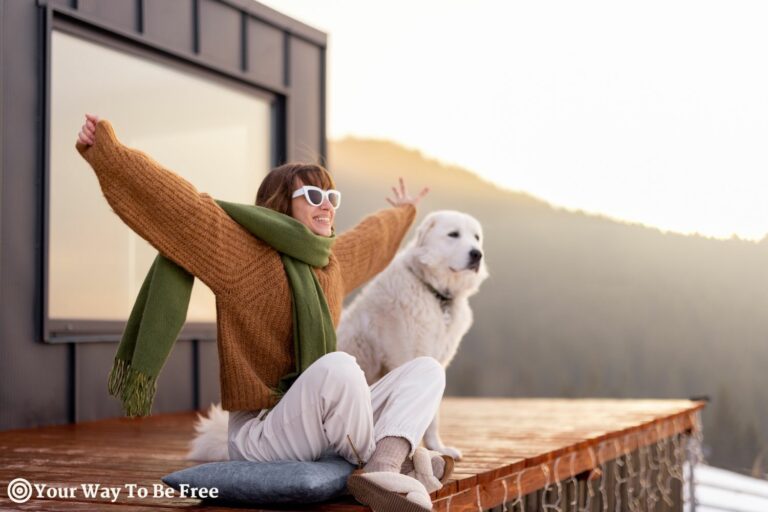 Woman sitting with dog on terrace of tiny house in the mountains enjoying beautiful sunrise landscape. Concept of small modern cabins for rest and escape to nature. morning winter routine